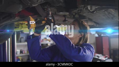 Arbeitsmechaniker in blauen Overalls mit Schlagschrauber unter dem Fahrzeug auf hydraulischem Hebezeug in der Werkstatt Stockfoto