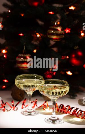 Zwei Gläser Sekt stehen auf einer weißen Oberfläche. Rotes Band und Tannenzapfen sitzen in der Nähe. Im Hintergrund leuchtet ein Weihnachtsbaum mit Lichtern und roten und goldenen Ornamenten. Stockfoto