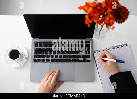 Minimalistischer Arbeitsplatz über Kopf: Laptop, Tasse Kaffee und Handschrift in einem Notizbuch mit warmem Herbstdekor. Perfekt für Fernarbeit, Planung und Fertigung Stockfoto