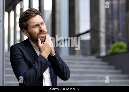 Geschäftsmann, der eine entzündete Wange vor dem Bürogebäude berührt, an Zahnschmerzen und Zahnfleischentzündungen leidet, dringend zahnärztliche Versorgung für starke Schmerzen benötigt Stockfoto