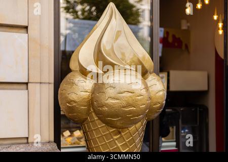 Vor einem Dessertladen steht eine große, auffällige Skulptur aus goldenem Eisbecher. Die Skulptur ist mit drei Schaufeln ausgestattet und zieht Passanten in A an Stockfoto