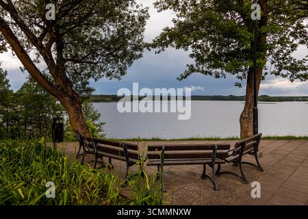 Park mit See, Blumenbeet und Bänken. See in einem bewaldeten Gebiet mit Pflanzen und Bäumen am Ufer unter bewölktem stürmischem Himmel Stockfoto