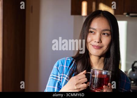 Asiatische Frau mit Glasbecher Tee in der Küche mit Edelstahl-Wasserkocher, Kopierraum Stockfoto