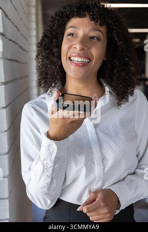 afroamerikanerin in weißer Bluse und schwarzer Hose mit Telefon zum Mund im Bürokorridor Stockfoto