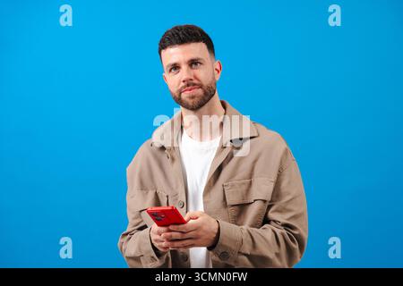 Junger bärtiger Mann, der ein rotes Smartphone auf blauem Hintergrund hält Stockfoto