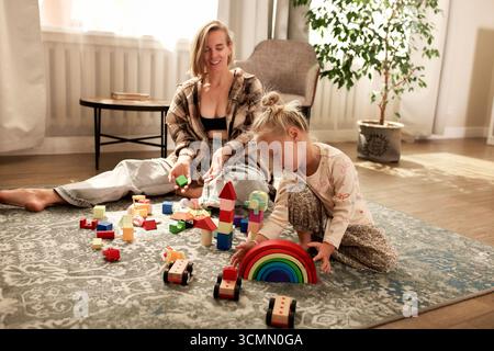 Ein fröhlicher Moment, in dem eine Mutter und ihr Kind in einem warmen, beleuchteten Zimmer zusammen mit hölzernen Bildspielzeugen auf einem Teppich spielen Stockfoto