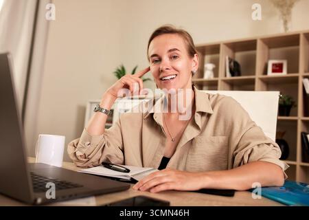 Lächelnde Frau, die an einem Schreibtisch sitzt und an Fernarbeit beteiligt ist. Der Arbeitsbereich verfügt über wichtige Gegenstände wie einen Laptop, ein Notizblock und eine Tasse, die für einen angenehmen Komfort sorgen Stockfoto