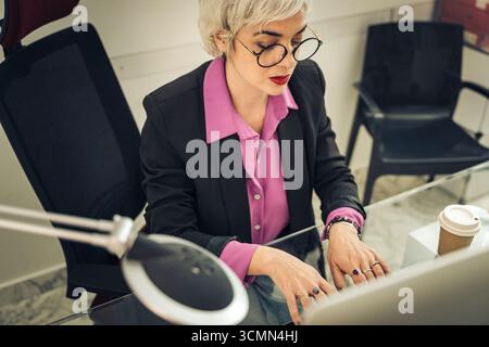 Aus einem hohen Blickwinkel konzentriert sich eine stilvolle Geschäftsfrau mit kurzen grauen Haaren und einer Brille auf ihre Arbeit, indem sie während einer Co-Party am Computer an einem Glasschreibtisch schreibt Stockfoto