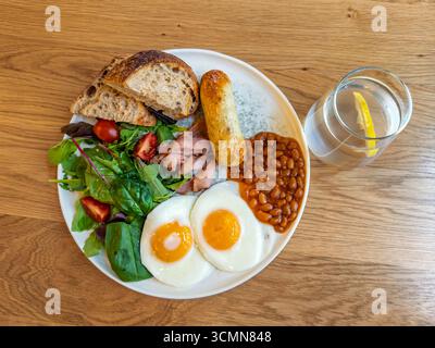 Komplettes englisches Frühstück mit Spiegeleiern, Speck, gebackenen Bohnen, Würstchen und frischem Salat auf weißem Teller mit Wasser, Blick von oben. Herzhafter Frühstücksteller Stockfoto