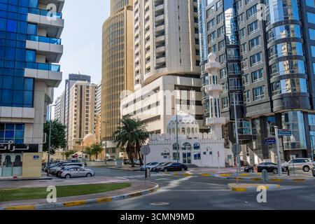 Städtische Kreuzung in Abu Dhabi, VAE, mit modernen Hochhäusern, die an klaren Tagen eine traditionelle weiße Moschee mit Kuppel und Minarett umgeben Stockfoto
