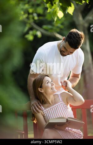 Junger Mann überraschte eine hübsche Freundin von hinten, während sie auf einer Bank saß und ein Buch las Stockfoto