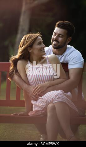 Die junge Frau, die auf der Bank sitzt, blickt glücklich auf einen schönen Freund zurück, während er sie umarmt Stockfoto
