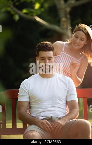 Junger Mann, der friedlich auf einer Bank sitzt, während die Freundin von hinten auf ihn zugeht Stockfoto