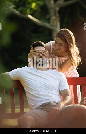 Junger Mann, der im Park von seiner schönen rothaarigen Freundin überrascht wird, deckt sie glücklich seine Augen hinter der Bank Stockfoto