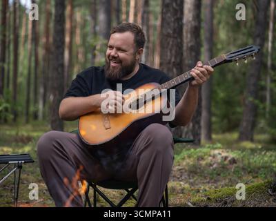 Bärtiger Musiker lächelt und spielt Akustikgitarre, während er in einem Campingstuhl neben einem warmen Lagerfeuer sitzt, umgeben von der ruhigen Schönheit des Fo Stockfoto