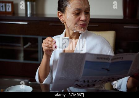 Eine ruhige Morgenszene mit einer Frau in einer Gesichtsmaske, Kaffee trinken und eine Zeitung lesen. Dieses Bild steht für Entspannung, Selbstfürsorge und Stockfoto