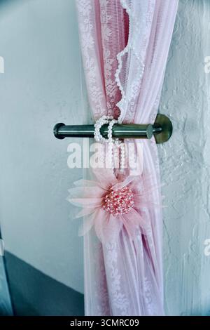 Pinkfarbener Spitzenvorhang mit Perlstickchen und perlenbesetzter Blume auf einem Metallstab Stockfoto