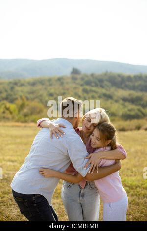 Eine Familie teilt sich eine fröhliche Umarmung in einem sonnigen Feld mit üppigem Grün und sanften Bergen im Hintergrund. Die Szene fängt Wärme und Zweisamkeit ein Stockfoto