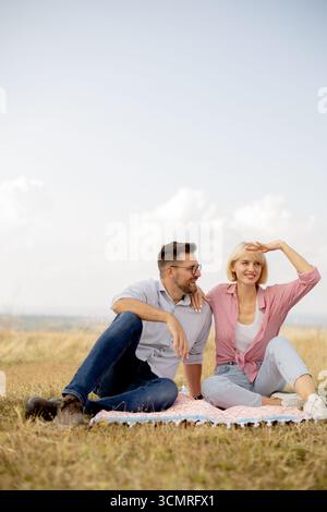 Ein junges Paar sitzt auf einer farbenfrohen Decke auf einem grasbewachsenen Gebiet. Sie lächeln und entspannen sich zusammen, genießen einen sonnigen Tag unter dem klaren blauen Himmel umgeben von Stockfoto