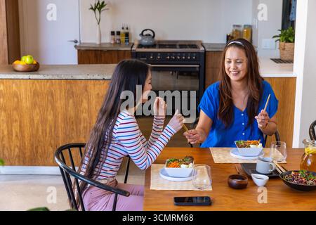 Asiatische Mutter und Tochter gehen Essstäbchen, während sie mit Reisschalen am Esstisch sitzen Stockfoto