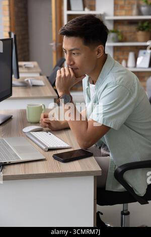 Asiatischer männlicher Büroangestellter, der auf einem Laptop mit Smartphone am Holzschreibtisch im Büro tippt Stockfoto