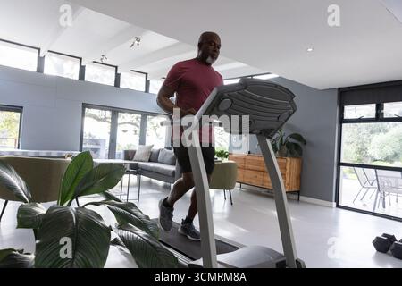 Rannter afroamerikaner, der zu Hause auf dem Laufband läuft und ein Hemd in der Nähe der Hantelfabrik trägt Stockfoto