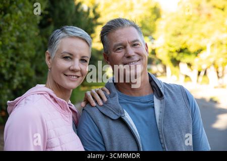 Seniorenpaar steht auf einem gepflasterten Wohnweg in Steppjacken und Reißverschlussjacken unter Sonnenlicht Stockfoto