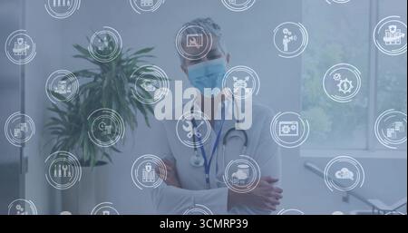 Stehender Arzt, der die Arme kreuzt, mit chirurgischer Maske und Stethoskop in der Klinik, mit digitalen Ikonen Stockfoto