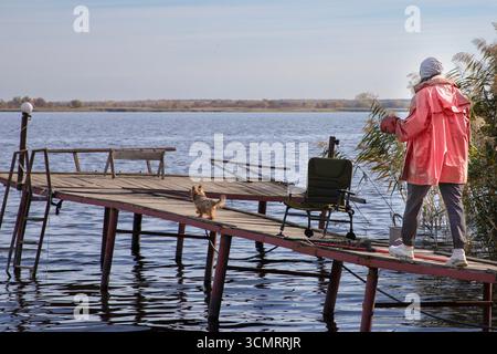 Rückansicht einer Person in hellem Jackett mit einem kleinen Hund, der im Herbst auf dem Pier am See steht. Konzept der Freizeit, Reise, Gesellschaft und Outdoor-lifestyl Stockfoto