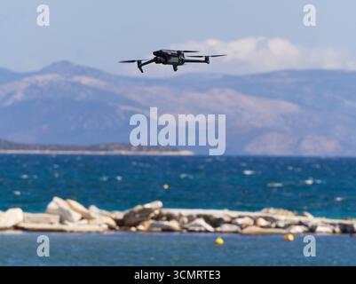 Schwarze Drohne schwebt über blauem Meereswasser mit felsiger Küste und Bergkette im Hintergrund Stockfoto