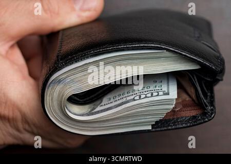 Die Hand eines Mannes hält eine schwarze Ledertasche mit einem Haufen 100-Dollar-Scheine, Banknoten, Bargeld, Nahaufnahme. Konzept für verlorenes oder gefundenes Wallet mit CA Stockfoto