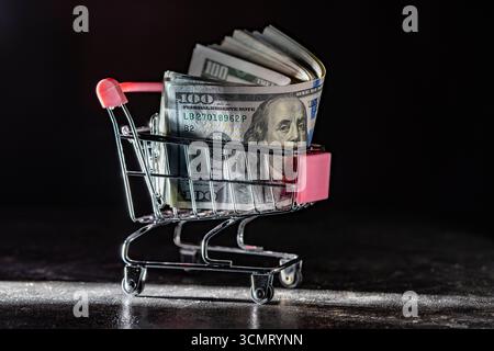 Warenkorb auf Rädern mit Dollarscheine auf dunklem Hintergrund, Nahaufnahme, Kopierraum. Finanz-, Geschäfts-, Einkaufs-, Ausgaben- und Vertriebskonzept Stockfoto