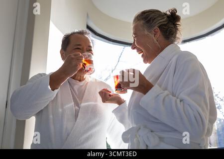 Verschiedene Seniorenpaare tragen Bademäntel mit Glasbechern, die Tee am Fenster teilen Stockfoto