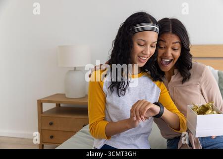 Verschiedene Mutter und Teenager-Tochter sitzen zu Hause auf dem Bett und halten weiße Geschenkbox Stockfoto