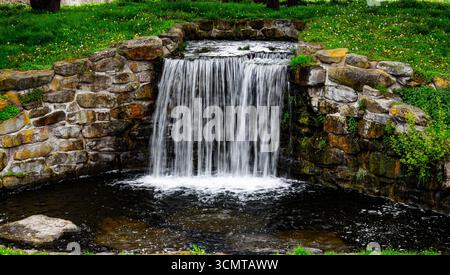 Wunderschöner künstlicher Wasserfall, der über verwitterte Steinmauern in landschaftlich gestaltetem Garten mit üppigem grünem Gras und natürlicher Felsformation stürzt Stockfoto