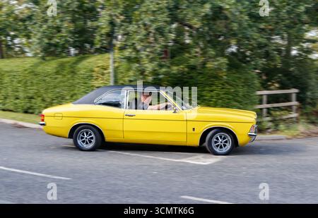 Gelber Vintage Ford Cortina Mk 3 mit schwarzem Vinyldach, der auf einer Landstraße mit Bäumen und Hecken fährt Stockfoto