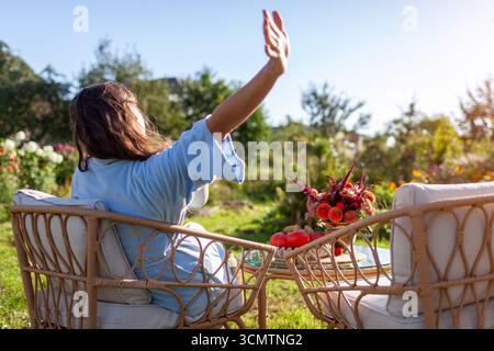 Rückansicht einer Frau, die sich morgens im Pyjama ausdehnt und sich auf Terrassenmöbeln im Herbstgarten entspannt. Kaffee zum Frühstück Stockfoto