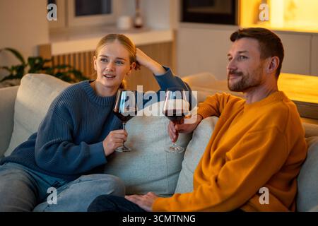 Glückliche junge Familie, die sich zu Hause auf dem Sofa entspannt und abends nach der Arbeit Wein trinkt, zusammen Fernsehen Stockfoto
