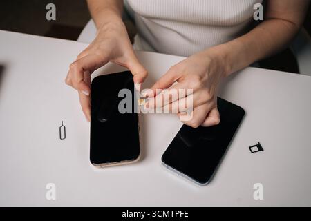 Nahaufnahme einer jungen Frau, die sim-Karte in ein neues Smartphone einsteckte, mit einem anderen Telefon und einem sim-Auswerferwerkzeug in der Nähe auf einem weißen Tisch, während eines mobilen Geräts Stockfoto