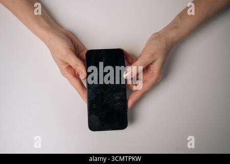 Hochwinkelansicht einer Frau, die den Displayschutz aus gebrochenem gehärtetem Glas vom Smartphone entfernt und eine gerissene Schutzfolie auf dem weißen Tisch freilegt. Stockfoto