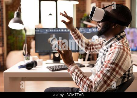 Schwarzer, männlicher Ingenieur, der ein VR-Headset trägt und die Hände in 3D-Simulation mit Gesten bewegt und das entwickelte Klemmmodell analysiert. Immersive Technologie ermöglicht interaktive technische Prüfungen und detaillierte Produktprüfungen. Stockfoto