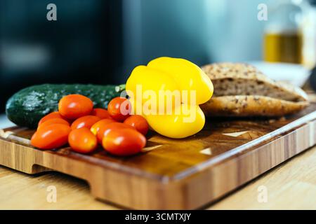 Bunte Auswahl an frischem Gemüse auf hölzernem Schneidebrett, mit gelbem Paprika, Kirschtomaten, Gurke und handwerklichem Brot, perfekt Stockfoto