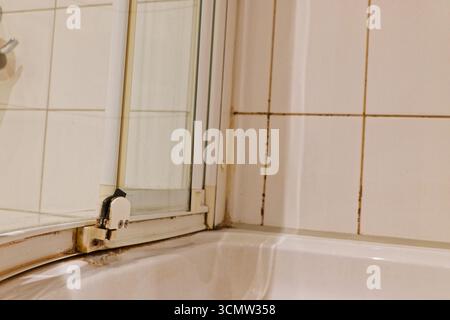 Weiße Fliesen säumen das Badezimmer, während eine Schiebetür entlang einer Metallschiene verläuft. Die Piste ist schwarz mit Schmutz und der Fugenmörtel zeigt gelben und dunklen Schimmel, fo Stockfoto