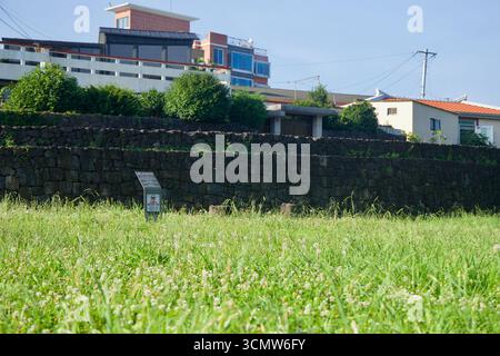 Steinmauern aus der historischen Seogwi Festung ragen über einem grasbewachsenen Feld mit einem Informationsschild und nahe gelegenen Häusern im Zentrum von Seogwipo. Stockfoto