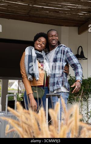 afroamerikanisches Paar, das auf der Veranda steht und lächelt mit Koffern, gestreiftem Schal, grüner Gürteltasche Stockfoto