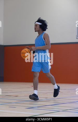 afroamerikanischer Mann in Uniform, weißes Stirnband, das Basketball über die Spielfelder des Fitnessstudios dribbelt Stockfoto