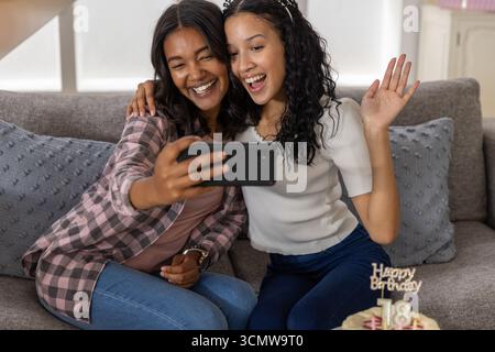 Vielfältige Mutter und Tochter im Teenageralter machen zu Hause Selfie auf dem Sofa mit Smartphone und Geburtstagskuchen Stockfoto