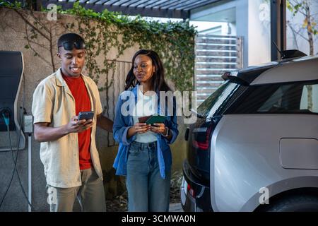 Afroamerikanische Freunde überprüfen Smartphone mit Karten unter Pergola am Elektroauto-Ladegerät Stockfoto