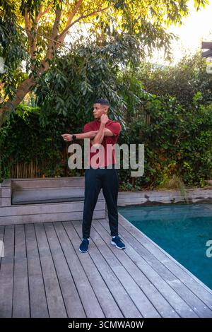 Afroamerikanischer Mann, der sich auf Holzterrasse neben dem Swimmingpool im Hinterhof mit Smartwatch ausdehnt Stockfoto