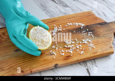 Nahaufnahme der Hand in einem schützenden Gummihandschuh, der Holzschneidebrett mit halber Zitrone und grobem Salz schrubbt. Konzept des natürlichen umweltfreundlichen NO Stockfoto